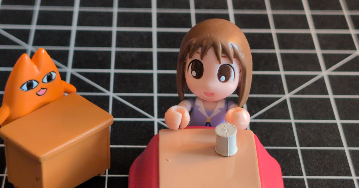 An Osaka toy from Azumanga Daioh sitting under a kotatsu with a strange cat at a desk next to her.